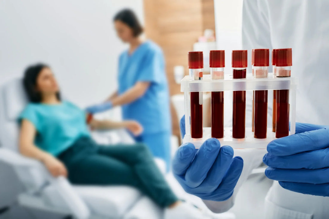 Gloved healthcare worker holding a rack of blood samples, with a patient and nurse in the background.