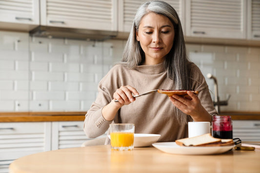 Grey-haired woman spreading jam on toast at breakfast, accompanying advice on coeliac disease later in life.