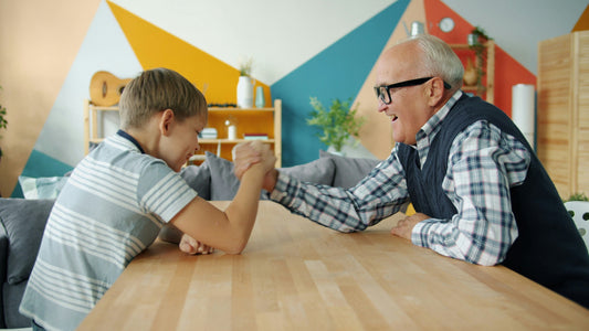 A grandfather and his grandson smile and laugh while arm-wrestling across a table.