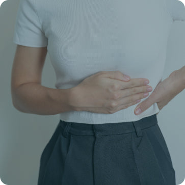 Person wearing a light gray t-shirt and dark gray pleated pants, holding their stomach, against a plain background